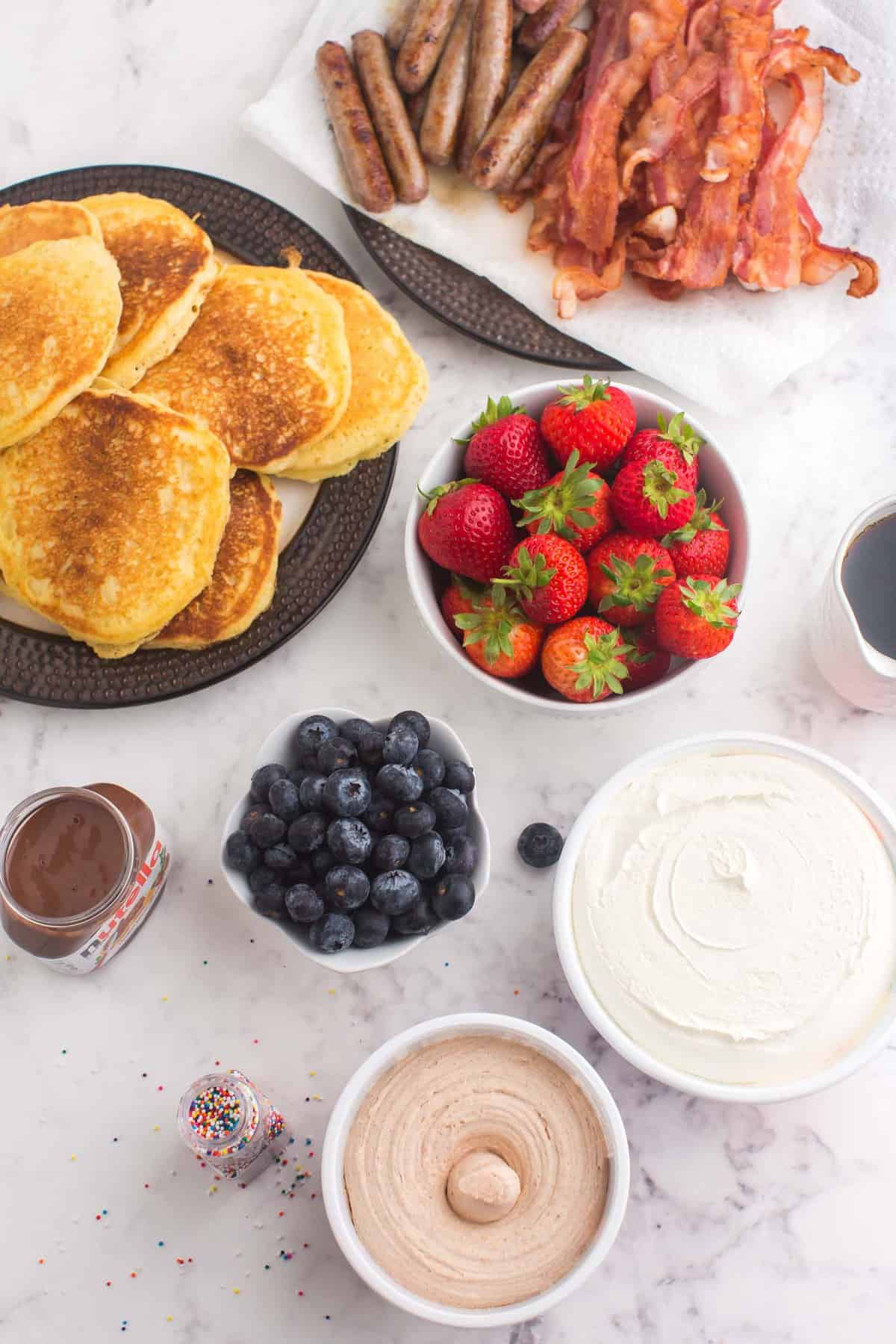 Ingredientes en cuencos y platos separados, panqueques, frutas, coberturas y carnes para el desayuno.