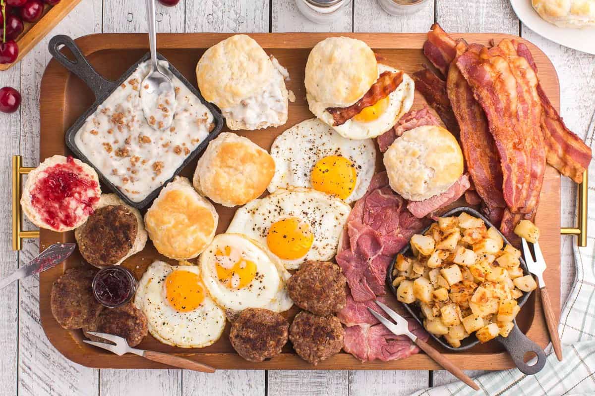 Huevos, tocino, papas, panecillos, salsa, mermelada y más en una gran tabla de madera rectangular.