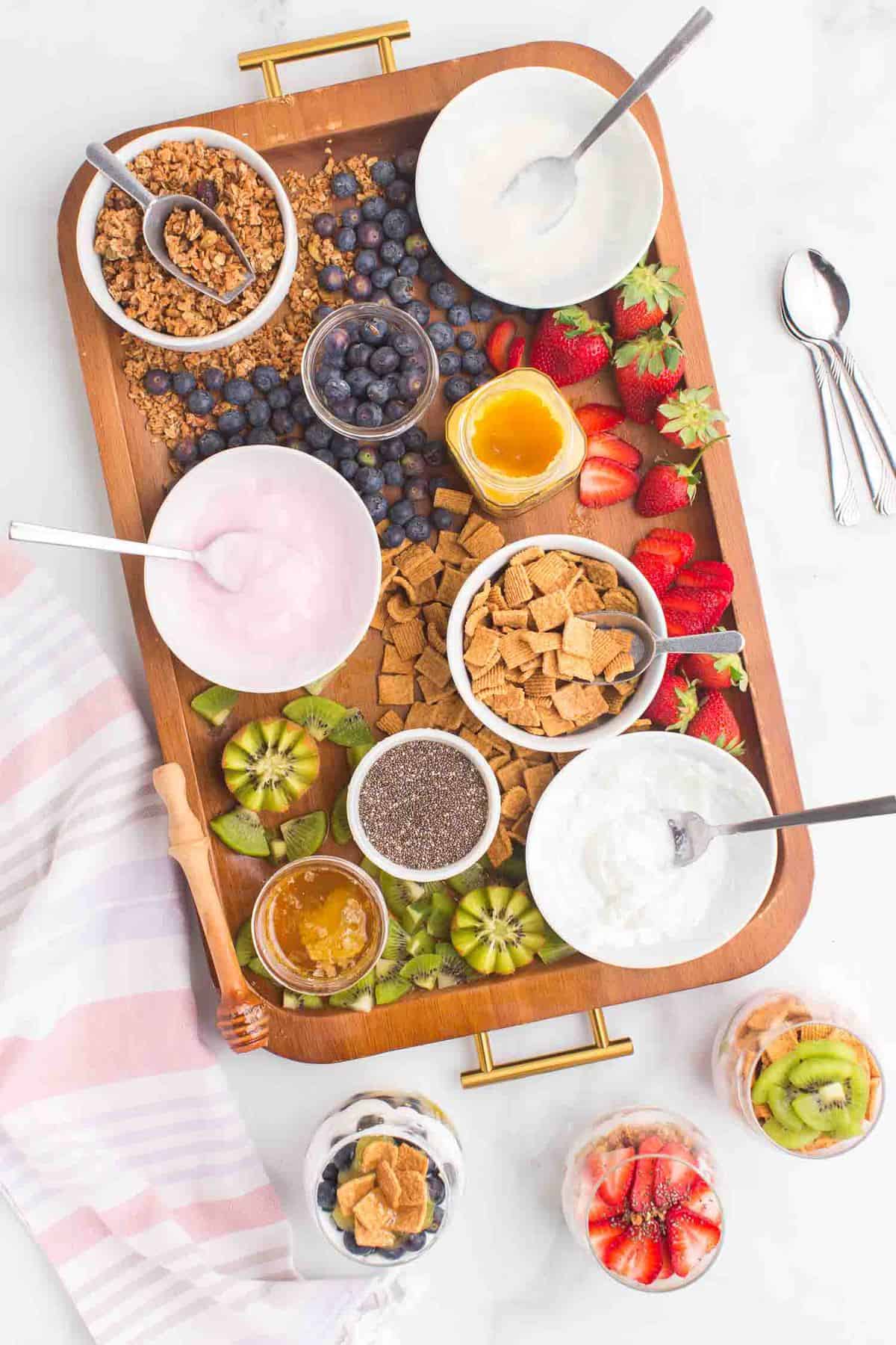 Yogurt y ingredientes dispuestos en una bandeja, tres parfaits al lado.