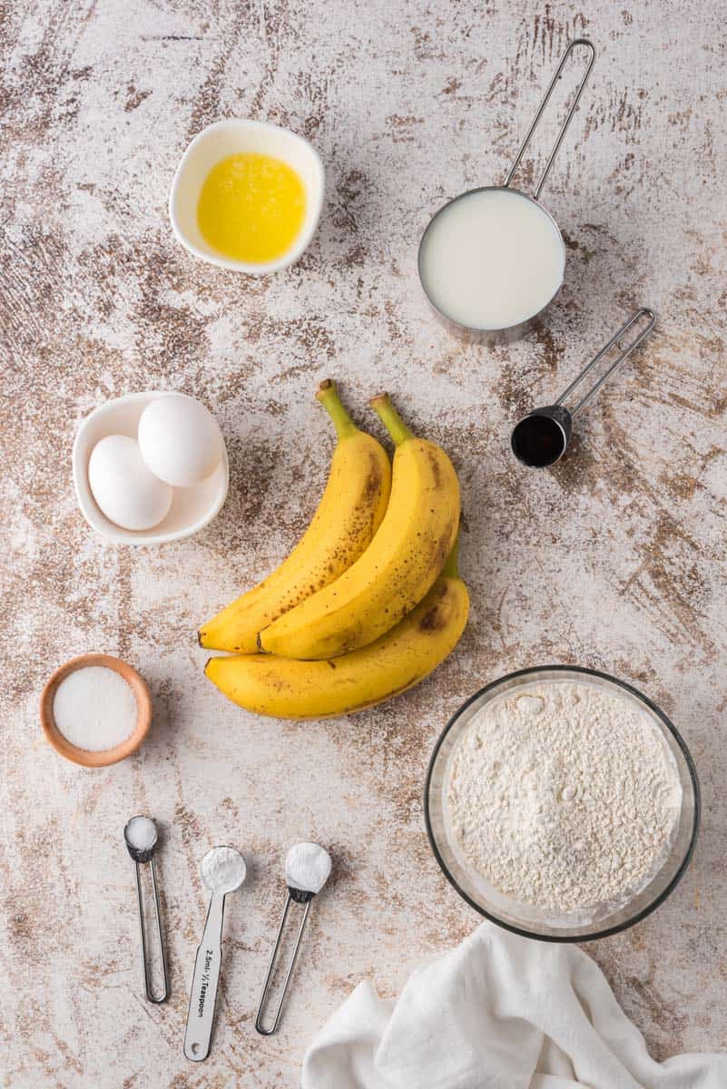 Ingredientes sobre una superficie marrón y blanca: harinas, azúcares, plátanos maduros, huevos, aceite, extracto de vainilla.