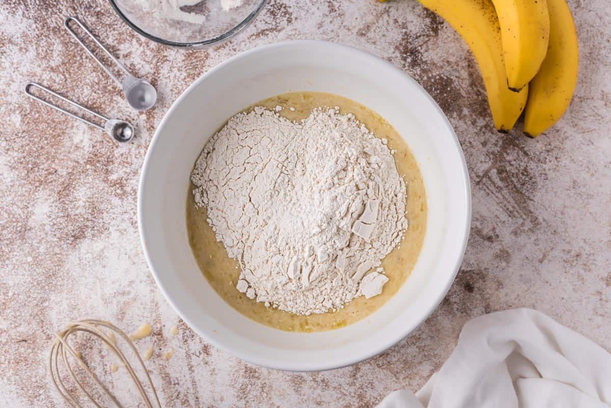 Ingredientes húmedos y secos en un tazón blanco. También se ven plátanos.