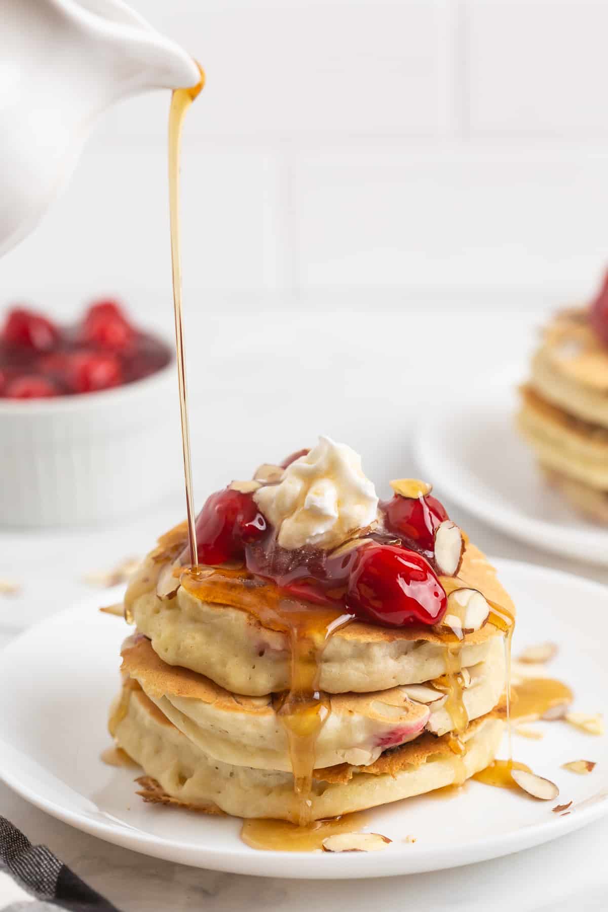 Cerezas y crema batida sobre una pila de panqueques, siendo bañada con jarabe.