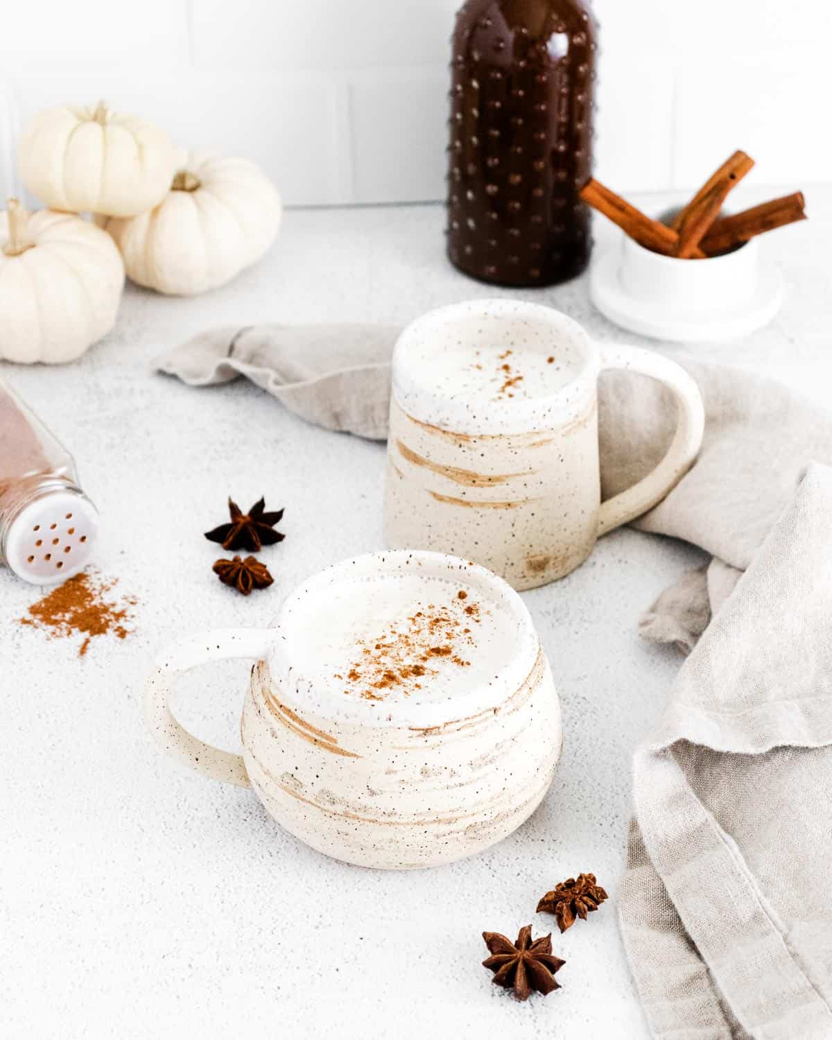 Latte de especias de calabaza en tazas de tonos tierra y blanco. También se muestran especias.
