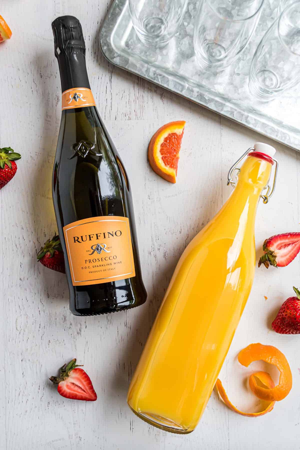 Una botella de prosecco y una botella de jugo de naranja, colocadas planas sobre una superficie gris claro.