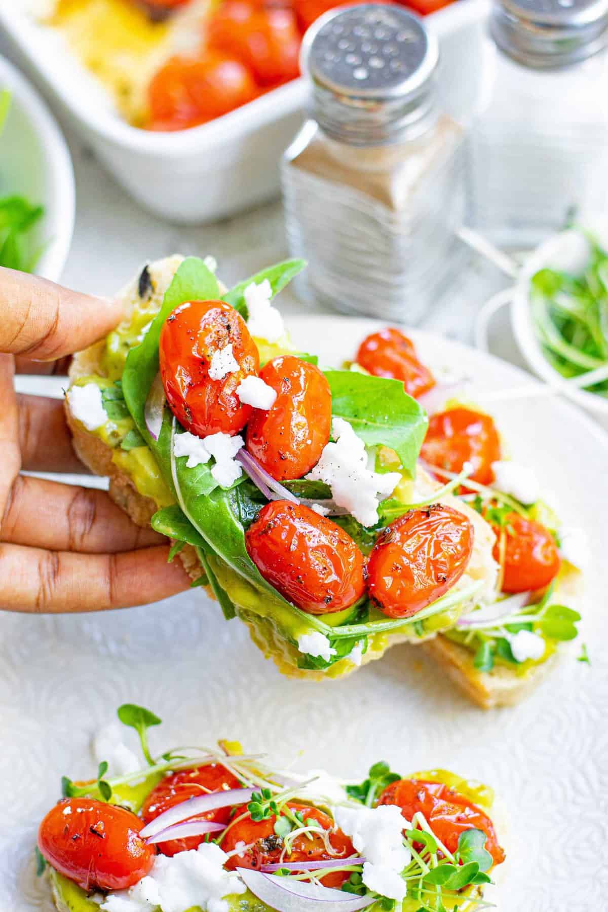 Mano sosteniendo una rebanada de pan cubierta con aguacate, tomate, feta y hojas verdes.