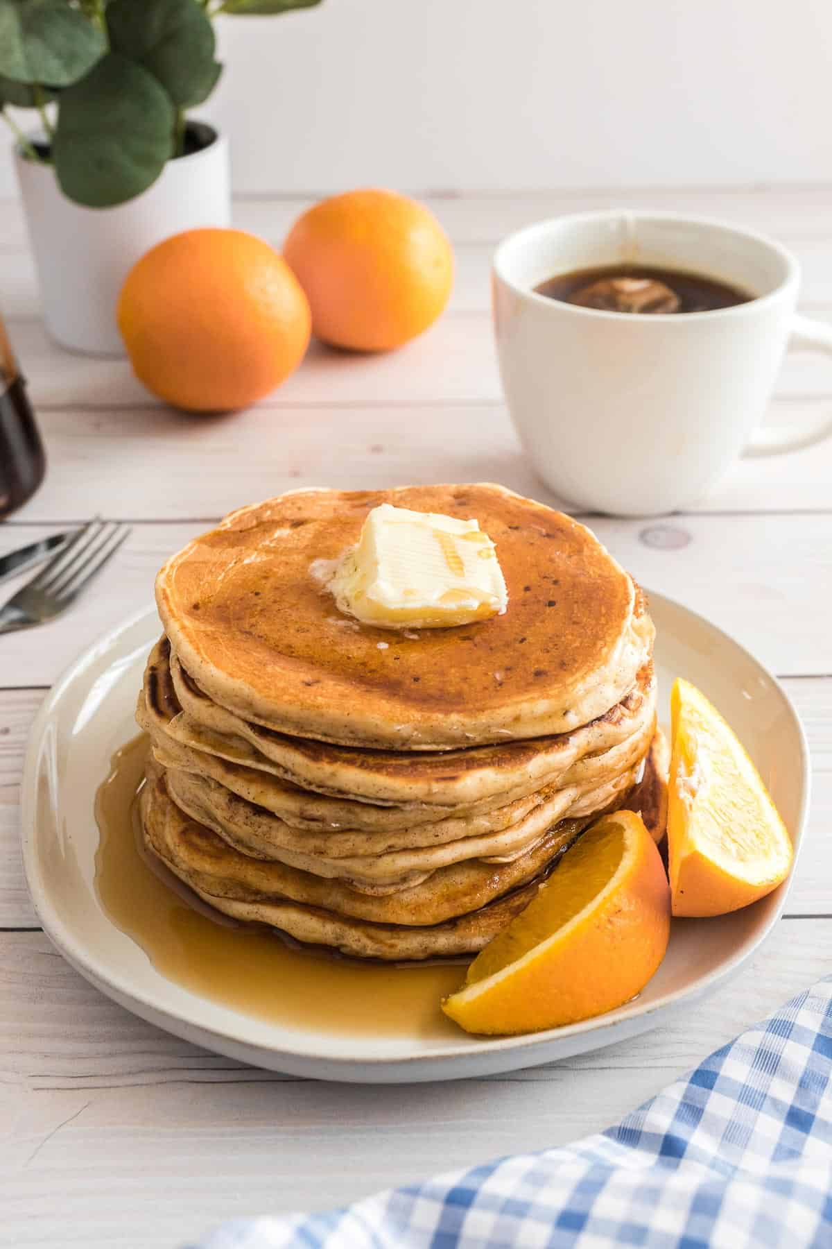 Panqueques y sirope en un plato con gajos de naranja, café y naranjas en el fondo.