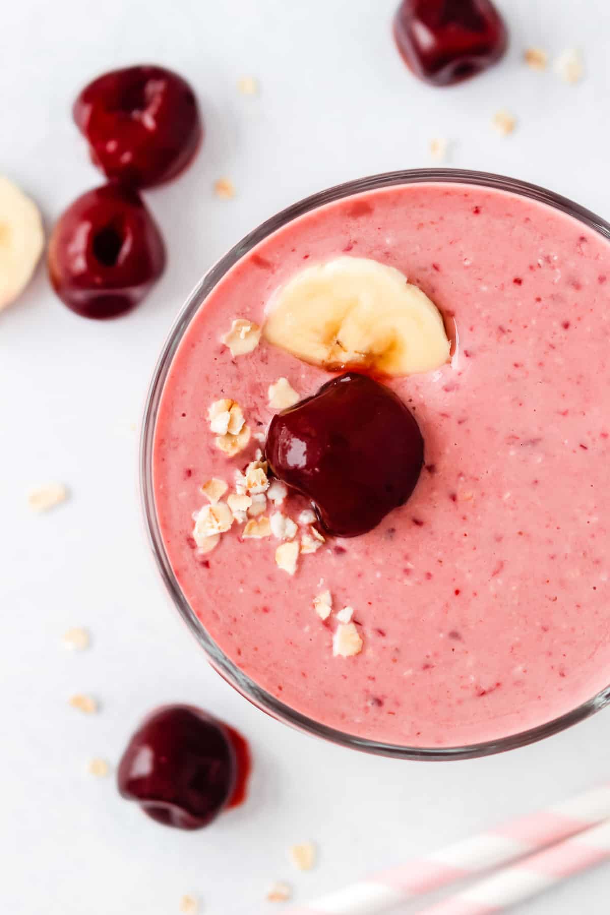 Vista aérea de un batido de cereza de color rosa claro, decorado con una cereza, una rodaja de plátano y avena.