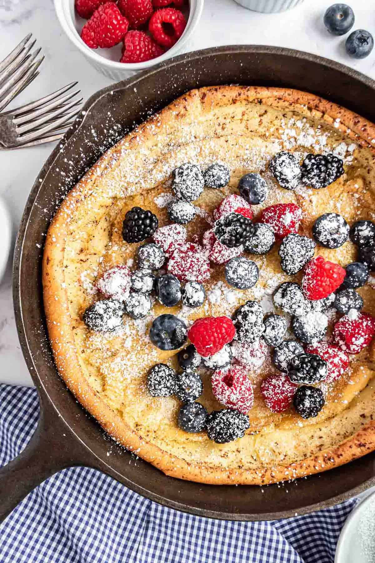 Panqueque Dutch baby en una sartén negra cubierto con bayas y azúcar en polvo.