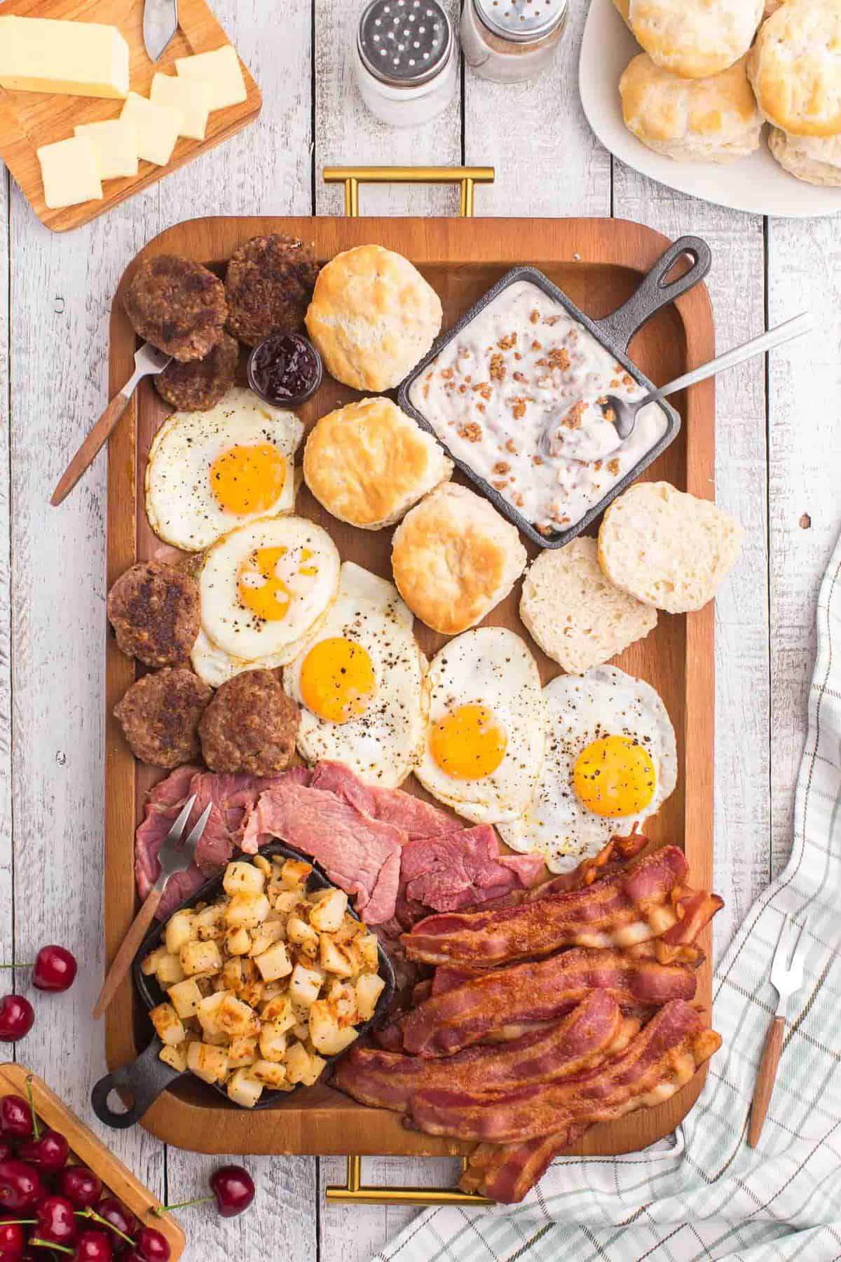 Vista aérea de carne, huevos, salchichas, papas y panecillos en una gran bandeja de madera.