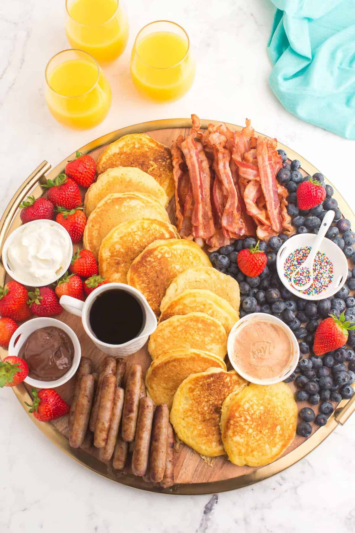Vista aérea de un desayuno dispuesto en una tabla, incluyendo panqueques, tocino, salchichas y frutas.