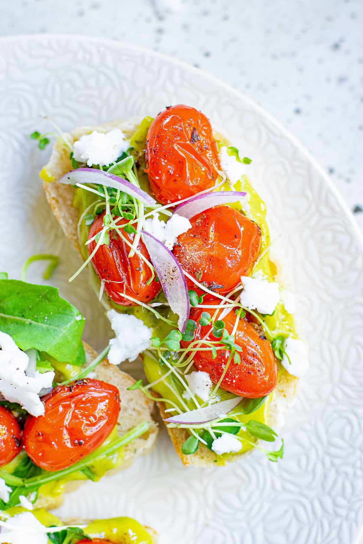 Vista aérea de una pequeña rebanada de pan cubierta con guacamole, tomates asados, hojas verdes y feta.