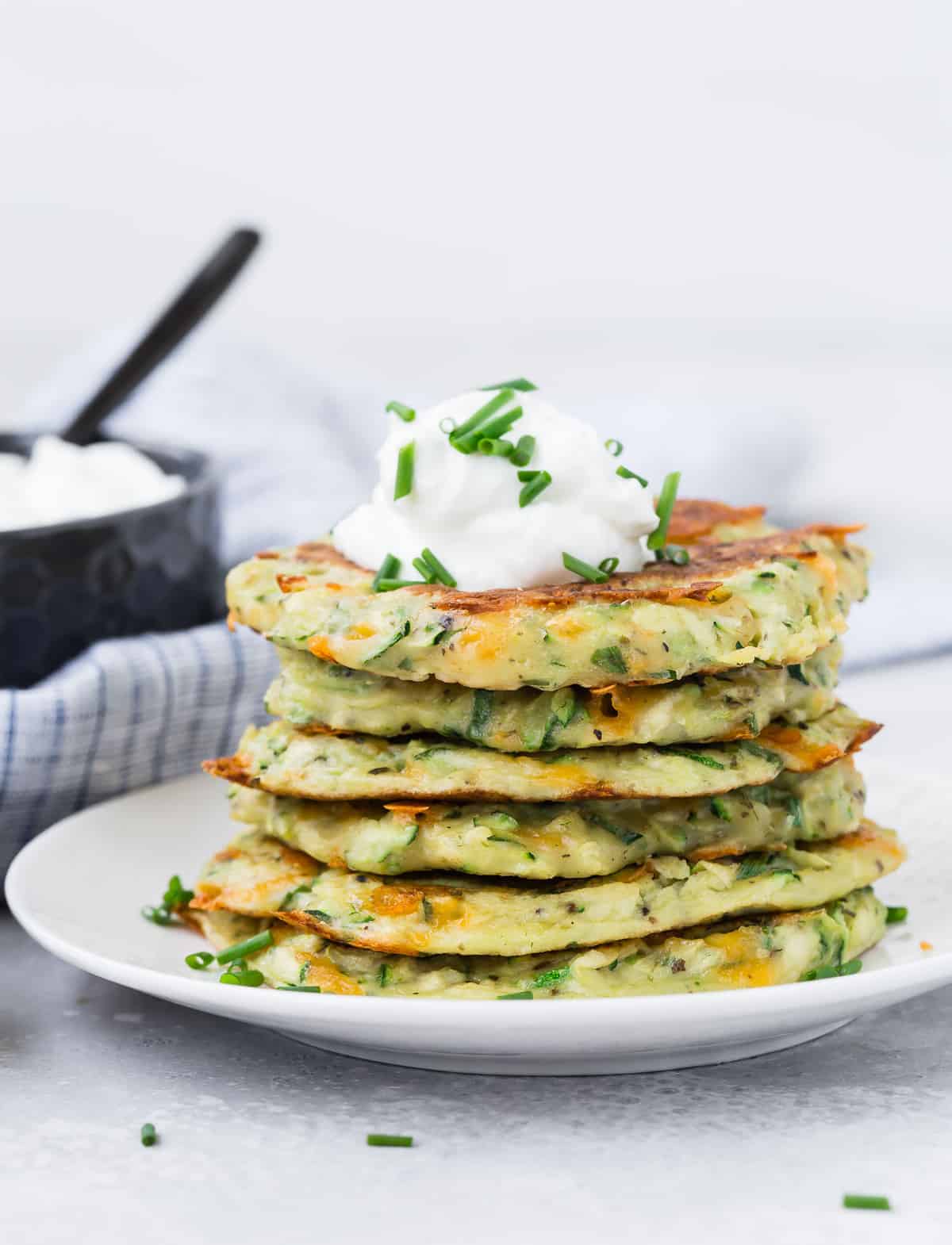 Una pila de panqueques de calabacín en un plato blanco, cubiertos con crema agria y cebollino.