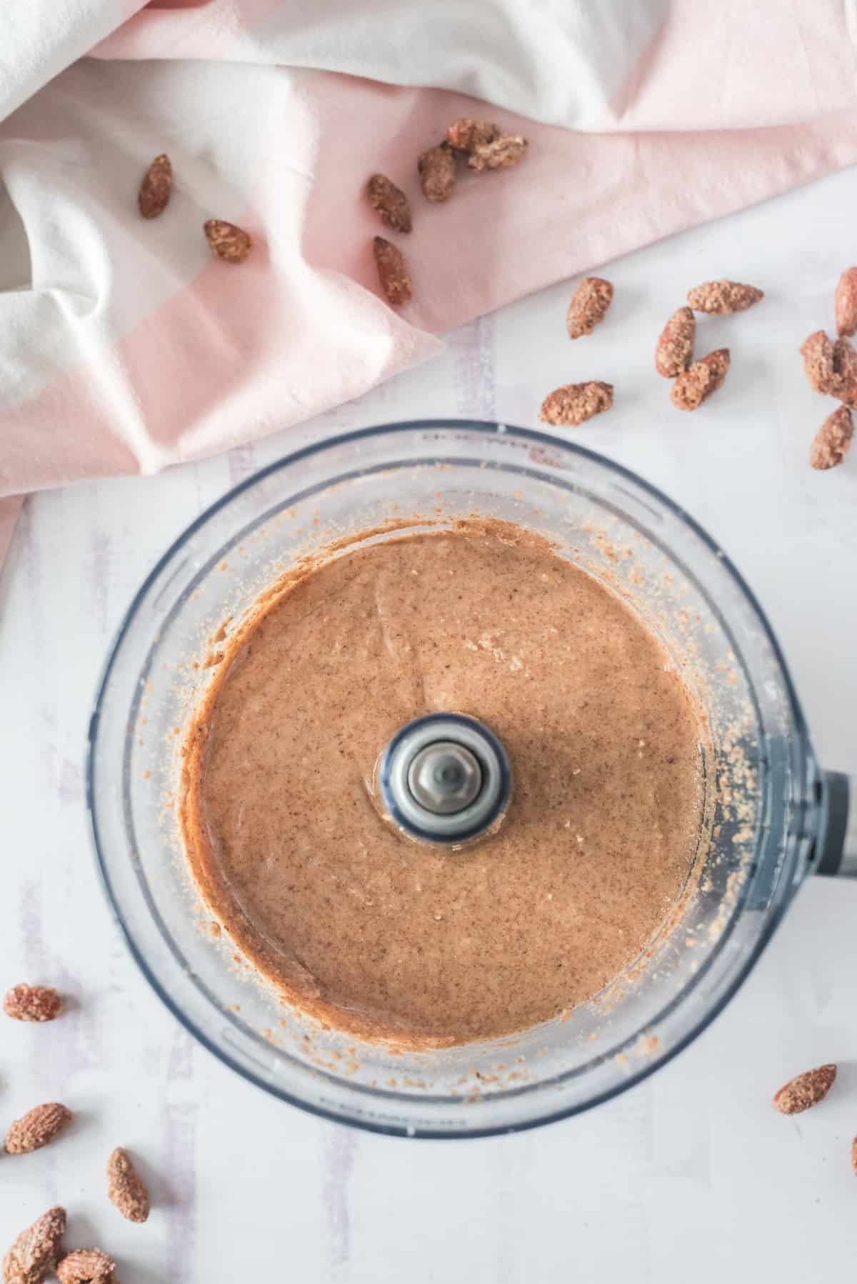 Mantequilla de almendra cremosa en un procesador de alimentos, con almendras esparcidas cerca.